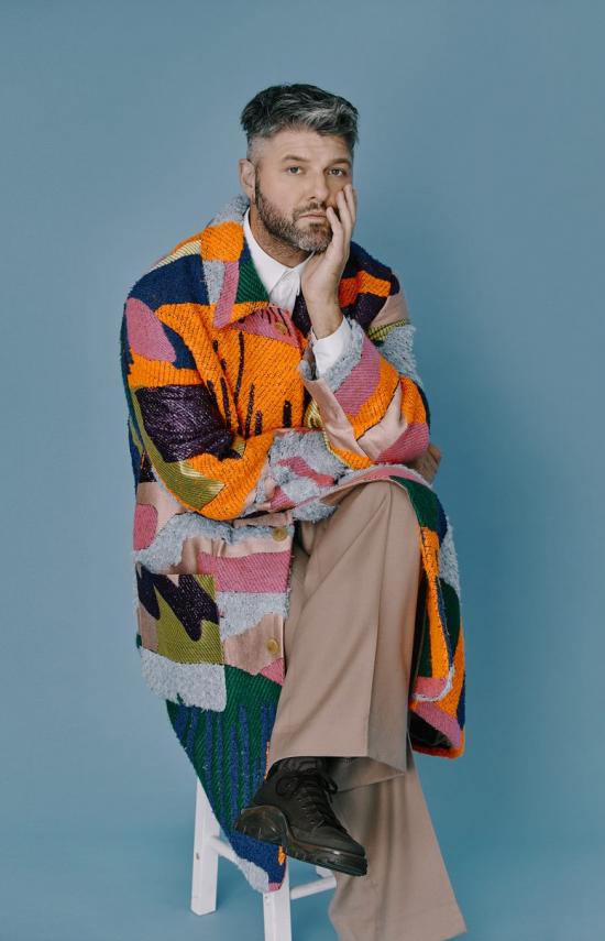 Photo de l'artiste Pierre Lapointe assis, la main gauche sur le visage, les jambes croisées avec une veste multicolores