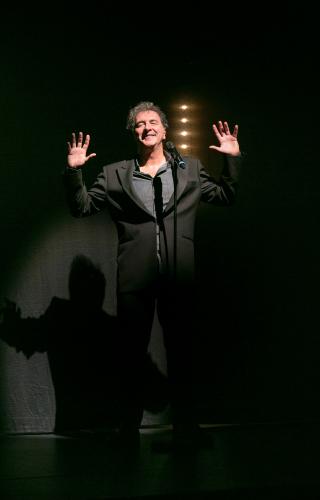 François Morel sur scène derrière un micro en costume noir, les mains levées